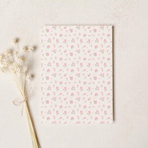 Op de afbeelding: Een rechthoekig notitieblok met een bloemenpatroon van kleine roze bloemen op een cr&egrave;mekleurige achtergrond. Een bosje gedroogde witte bloemen, vastgebonden met touw, bevindt zich links van het notitieblok.