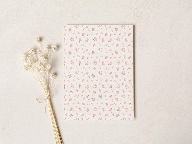 Op de afbeelding: Een rechthoekig notitieblok met een bloemenpatroon van kleine roze bloemen op een cr&egrave;mekleurige achtergrond. Een bosje gedroogde witte bloemen, vastgebonden met touw, bevindt zich links van het notitieblok.