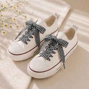 May include: A pair of white canvas sneakers with black and white gingham ribbon laces. The shoes have a ribbed toe cap and a thin burgundy stripe along the sole. The sneakers are set on a white surface.