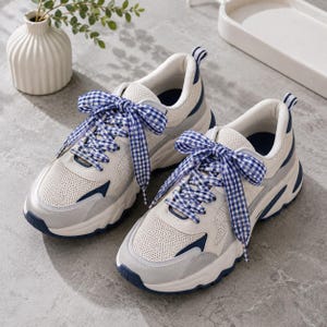 May include: A pair of cream-colored athletic shoes with mesh panels, gray accents, and navy blue details. The shoes feature thick white soles and are tied with blue and white gingham ribbon laces. The shoes are designed for comfort and style.