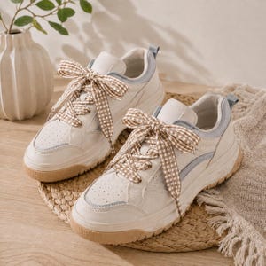 Puede incluir: Un par de zapatillas blancas con detalles en azul claro y beige. Los zapatos tienen suelas beige y están atados con cintas de cuadros marrones y blancas. Las zapatillas están sobre una estera tejida.