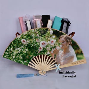 May include: A decorative fan with a photo of a woman smelling flowers, with the text "Sienna & Dominic". The fan has a wooden handle and a blue tassel. The words "Individually Packaged" are also visible.