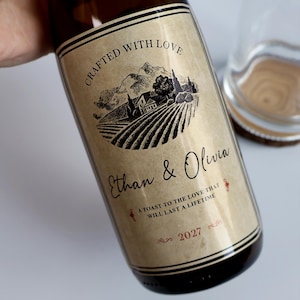 May include: A glass wine bottle with a beige label featuring a black ink illustration of a vineyard and mountains. The text reads Crafted with Love, Ethan & Olivia, A toast to the love that will last a lifetime, and the year 2027 in red ink.