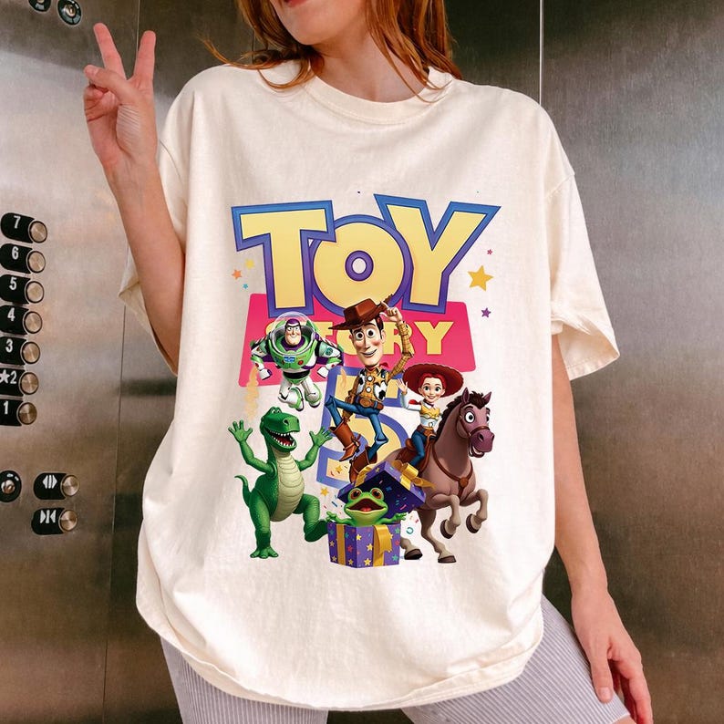 Puede incluir: Una camiseta de manga corta de color crema con un gran estampado gr&aacute;fico de los personajes de Toy Story, incluyendo a Woody, Buzz Lightyear, Jessie, Rex, Bullseye y un alien&iacute;gena verde en una caja de regalo, con el texto TOY STORY en letras amarillas.