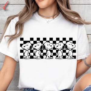 Op de afbeelding: Wit T-shirt met een zwart-wit afbeelding van Snoopy en een dambordpatroon. De cartoon Snoopy wordt vijf keer herhaald over het shirt. Het T-shirt heeft een klassieke ronde hals.
