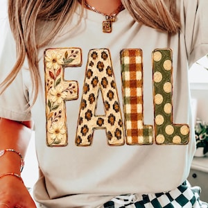 May include: A beige t-shirt featuring the word FALL in large block letters. The F has a floral pattern, the A has a leopard print, the first L has a brown and green plaid pattern, and the second L has a green background with gold polka dots.