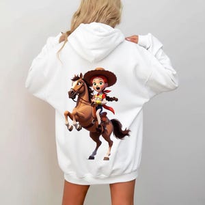 Puede incluir: Una sudadera con capucha blanca que presenta un estampado gr&aacute;fico en la espalda. El dise&ntilde;o muestra a una vaquera de dibujos animados con cabello rojo, sombrero marr&oacute;n y pa&ntilde;uelo rojo, montando un caballo marr&oacute;n. Lleva una camisa amarilla.