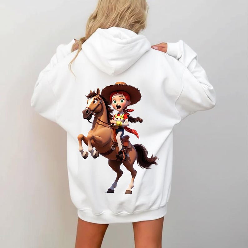 Puede incluir: Una sudadera con capucha blanca que presenta un estampado gr&aacute;fico en la espalda. El dise&ntilde;o muestra a una vaquera de dibujos animados con cabello rojo, sombrero marr&oacute;n y pa&ntilde;uelo rojo, montando un caballo marr&oacute;n. Lleva una camisa amarilla.