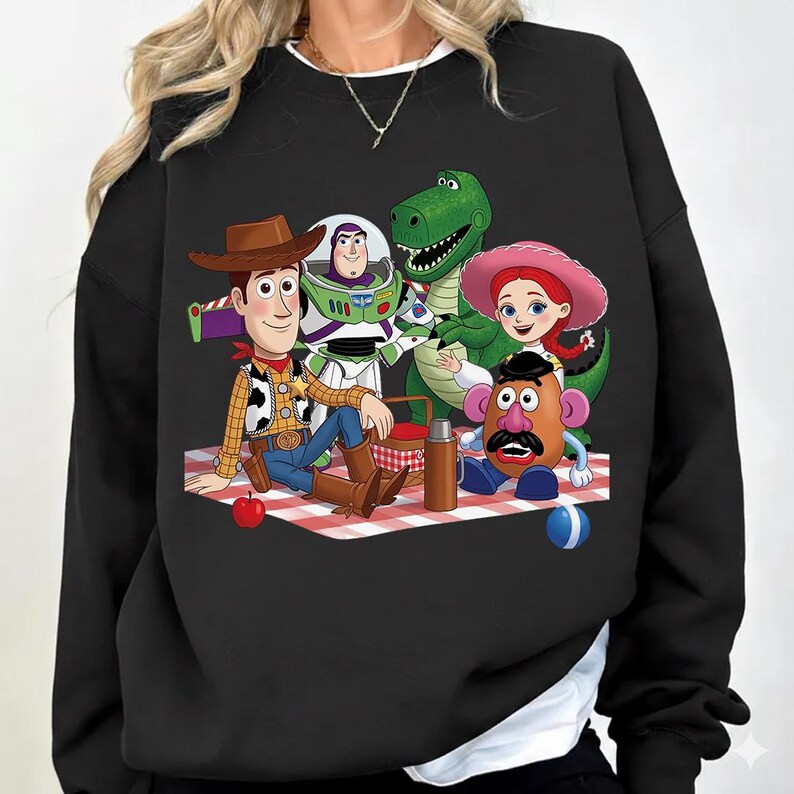 Puede incluir: Una sudadera negra de manga larga con un estampado gr&aacute;fico de personajes de Toy Story, incluidos Woody, Buzz Lightyear, Rex, Jessie y el Sr. Potato Head, sentados sobre un mantel de picnic a cuadros rojos y blancos con una manzana roja y una pelota azul.