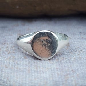 May include: A silver signet ring with a smooth, oval face. The ring has a polished finish and a classic design. The ring is set against a neutral background, highlighting its simple elegance. A timeless piece of jewellery.