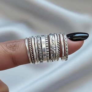 May include: A collection of silver rings with various designs, including twisted rope, floral patterns, and geometric shapes. The rings are stacked on a finger, showcasing their different textures and styles. The rings are made of a shiny metal.