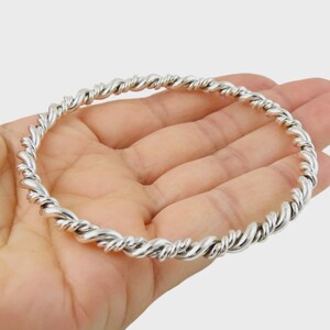Puede incluir: Un brazalete de cuerda retorcida de color plateado. El brazalete está hecho de un material metálico brillante y tiene un diseño trenzado texturizado. El brazalete se muestra en la mano de una persona.