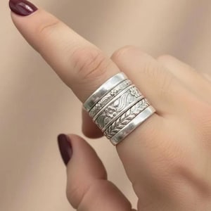 May include: A silver ring with multiple bands, each with a different embossed design. The designs include floral patterns and leaf motifs. The ring is worn on a finger, showcasing the intricate details of the jewellery.