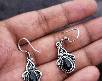 May include: A pair of silver-toned drop earrings with a teardrop-shaped black gemstone. The earrings have a decorative design around the gemstone and a hook closure. The earrings are displayed against a neutral background.