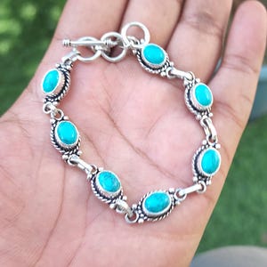 May include: Silver-toned bracelet featuring oval turquoise stones. The bracelet has a decorative, detailed setting around each stone and a toggle clasp. The bracelet is held in a hand against a blurred green background.