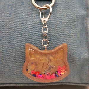 May include: A cat-shaped keychain with a silver clasp and chain. The clear resin pendant contains a silver cat charm, colorful heart-shaped confetti, and the word "ONE". The background is a light denim fabric.