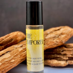 Puede incluir: Un frasco roll-on de vidrio transparente con tapa negra y etiqueta amarilla. La etiqueta dice "THE EMPORER" con texto adicional. El frasco está rodeado de trozos de madera marrón claro.