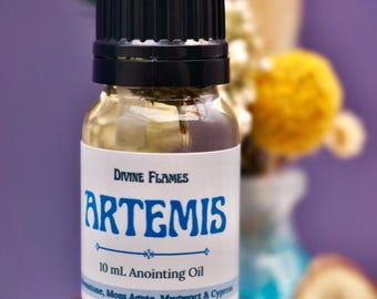 May include: A clear glass bottle of Artemis anointing oil with a black cap. The label reads "Divine Flames ARTEMIS 10 mL Anointing Oil". The bottle sits on a brown, textured surface. The background is blurred with purple and yellow accents.