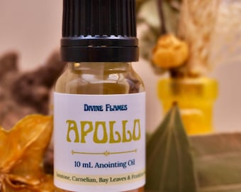 May include: A clear glass bottle with a black cap, labeled "APOLLO 10 mL Anointing Oil." The label has gold lettering and the words "Divine Flames." The bottle is surrounded by dried leaves and other natural elements.