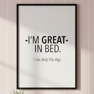 May include: A framed print with the text "-I'M GREAT- IN BED. I can sleep for days." The print has a white background and black text, framed in a black frame. A small potted plant sits below the frame.
