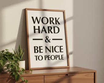 Puede incluir: Un cuadro enmarcado con el texto "WORK HARD & BE NICE TO PEOPLE" en letras negras. El marco es marrón y el fondo es blanquecino. El cuadro se exhibe sobre una cómoda de madera, con plantas en macetas blancas.