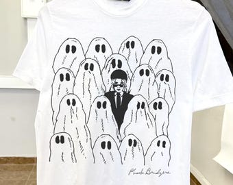 Op de afbeelding: Wit t-shirt met een zwart-wit afbeelding van een persoon in pak en zonnebril, omringd door spookfiguren. De tekst "Phoebs Bridgers" is eronder gedrukt.