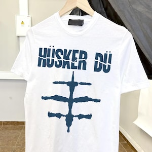 Op de afbeelding: Wit t-shirt met de tekst "HÜSKER DÜ" in donkerblauw, boven een donkerblauw abstract ontwerp. Het t-shirt hangt aan een houten hanger.