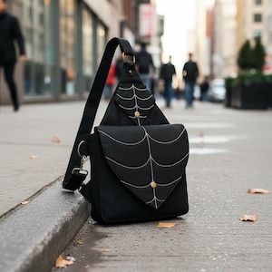 Könnte beinhalten: Schwarze Umhängetasche mit einem Spinnennetz-Design, das mit weißem Faden genäht ist. Die Tasche hat eine dreieckige obere Tasche und ein rechteckiges Hauptfach. Goldfarbene Hardware akzentuiert den Riemen und die Verschlüsse.