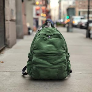 Puede incluir: Una mochila verde con múltiples bolsillos y un asa superior está sobre una acera. La mochila tiene un bolsillo frontal rectangular y dos bolsillos laterales. El fondo está borroso, mostrando una calle de la ciudad con edificios y coches.