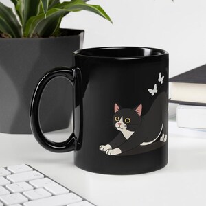 Puede incluir: Una taza de cerámica negra con una ilustración gráfica de un gato blanco y negro en posición de estiramiento. El gato tiene ojos amarillos y está situado bajo dos pequeñas mariposas blancas. La taza tiene un acabado brillante y asa.