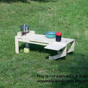 May include: A light-coloured wooden camping table with a built-in stove, a pot, a bowl, and a container. The table is set up on grass. The text on the image says: "This is not a product. It is an assembly guide with blueprints."