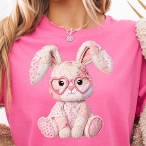 Puede incluir: Una camiseta rosa con un diseño de conejo de patchwork. El conejo lleva gafas en forma de corazón y está hecho de tela con estampados florales y de lunares en tonos rosa y crema. Las orejas del conejo están levantadas y tiene una expresión dulce.
