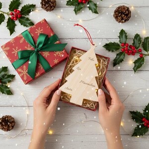 Könnte beinhalten: Ein hölzerner Weihnachtsbaumschmuck mit eingravierten Namen wird in einer roten Geschenkbox mit Stroh präsentiert. Eine rote Geschenkbox mit grüner Schleife befindet sich in der Nähe. Stechpalme, Tannenzapfen und Lichterketten schmücken die weiße Holzoberfläche.