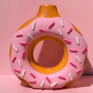 Könnte beinhalten: Eine Keramikvase in Donutform mit rosa Zuckerguss und bunten Streuseln. Die Vase hat ein Loch in der Mitte und einen braunen Boden und Hals. Der Hintergrund ist einfarbig rosa.