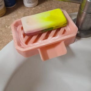 May include: A pink soap dish with a rectangular bar of soap. The soap is a gradient of yellow, white, and pink. The dish has a slotted design and a small tab on the front. The soap dish is sitting on the edge of a white sink.