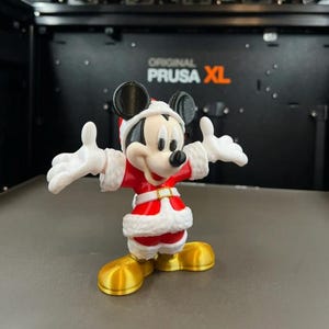 May include: A 3D-printed Mickey Mouse figurine in a Santa Claus outfit. The figure is red and white with gold shoes and a black face and ears. The background shows a black machine with the text "ORIGINAL PRUSA XL".
