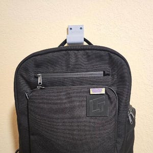 Pode incluir: Mochila preta com vários compartimentos com zíper e um bolso frontal. A mochila tem um tecido texturizado e um pequeno patch com o logotipo. Está pendurada em um gancho cinza.