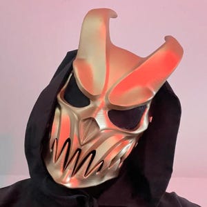 May include: A gold-colored mask with a horned design and sharp teeth, worn with a black hooded garment. The mask has a menacing appearance, with a detailed, metallic finish. The overall aesthetic suggests a fantasy or cosplay theme.