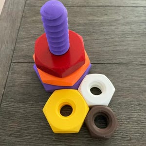 May include: A colorful wooden toy set featuring a purple screw and various hexagonal nuts in red, orange, yellow, white, and brown. The screw is stacked on top of the nuts, creating a playful and educational toy.
