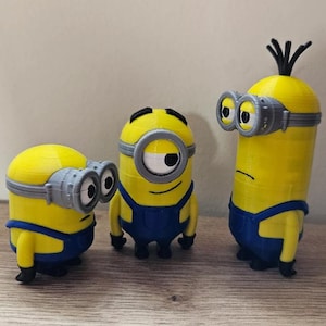 Può includere: Tre statuette gialle dei Minion con salopette blu e occhiali grigi. I Minion hanno espressioni e acconciature diverse. Uno ha un solo occhio, un altro ha un'acconciatura alta e il terzo ha due occhi.