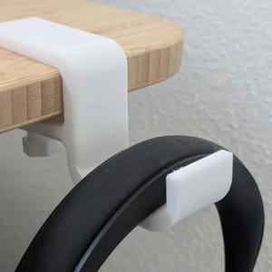 May include: A white headphone hanger is attached to a wooden desk. The hanger is designed to hold headphones and keep them organized. The desk is made of light brown wood.