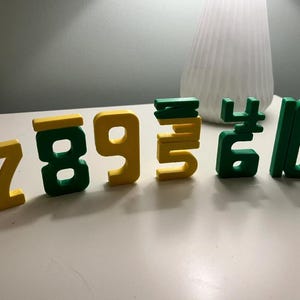 Op de afbeelding: Houten cijferblokken in geel en groen, gerangschikt op een wit oppervlak. De cijfers 7, 8, 9, 5, 4 en 10 zijn zichtbaar. De blokken zijn waarschijnlijk voor educatieve of decoratieve doeleinden.