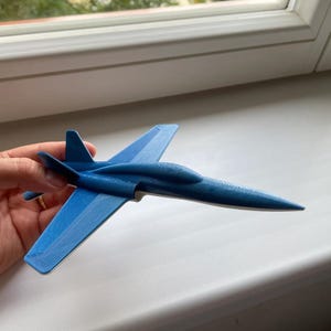 Puede incluir: Un modelo de avión azul con un diseño elegante. El avión de juguete tiene alas, cola y cabina. El avión se sostiene en una mano, y el fondo es una ventana y un alféizar blanco.