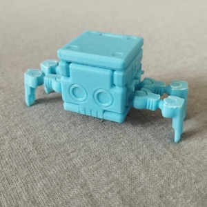 Op de afbeelding: Een lichtblauw, 3D-geprint robotspeelgoed met een doosvormig lichaam en gelede poten. De robot heeft twee cirkelvormige kenmerken aan de voorkant en een gestructureerd oppervlak. Het speelgoed is ontworpen om op een mechanisch insect te lijken.