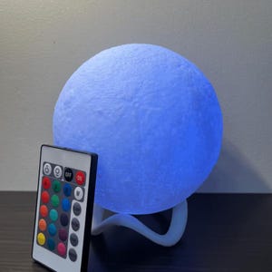 May include: A blue moon-shaped lamp with a textured surface, resting on a white stand. A remote control with color options and on/off buttons is positioned in front of the lamp. The lamp emits a soft blue glow.