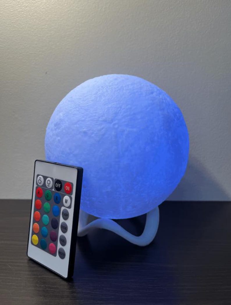 May include: A blue moon-shaped lamp with a textured surface, resting on a white stand. A remote control with color options and on/off buttons is positioned in front of the lamp. The lamp emits a soft blue glow.