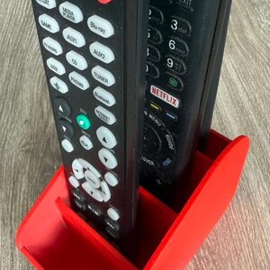 May include: A red 3D-printed remote control holder containing two black remotes. The remotes feature buttons such as "Power," "Menu," and "Netflix." The holder is designed to keep remotes organised and accessible.