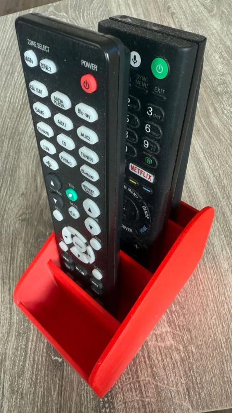 May include: A red 3D-printed remote control holder containing two black remotes. The remotes feature buttons such as "Power," "Menu," and "Netflix." The holder is designed to keep remotes organised and accessible.