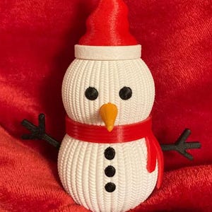 May include: A 3D-printed snowman figurine with a red hat, scarf, and arms. The snowman has black eyes and buttons, and an orange nose. The body is white with a textured pattern. A festive winter decoration.
