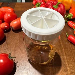 Puede incluir: Un tarro de cristal transparente con tapa blanca, lleno de líquido dorado. La tapa tiene seis compartimentos. Tomates rojos y pimientos de varios colores rodean el tarro sobre una superficie de madera.